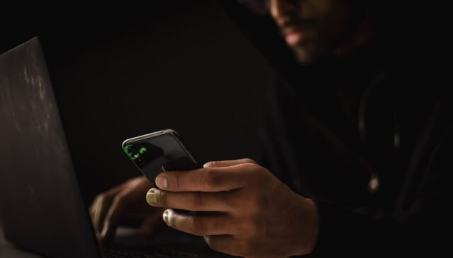 crop ethnic hacker with smartphone typing on laptop in dark room