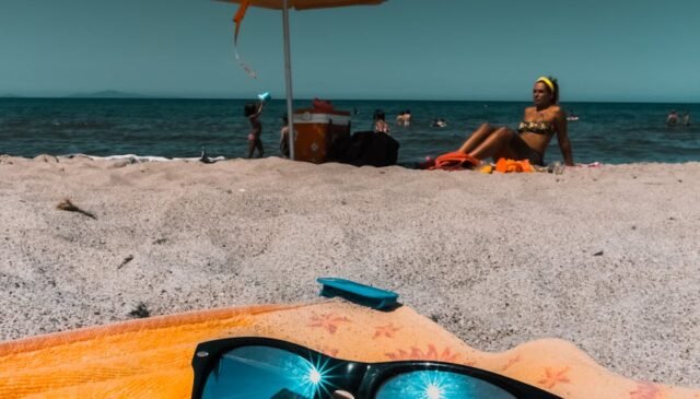 black framed sunglasses on towel with woman sitting on beach sand on background